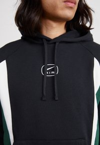 Musta huppari, jossa on valkoiset ja vihreät sivupaneelit. Siinä on nyöritettävä huppu ja pyöreä logo rinnassa. Pehmeää puuvillakangasta.