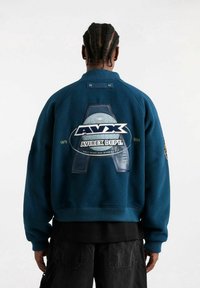 Man met gevlochten haar die een blauwe bomberjack draagt met een groot Avirex-logo en de tekst "Slechts enkelen zijn geboren om te vliegen" op de achterkant.