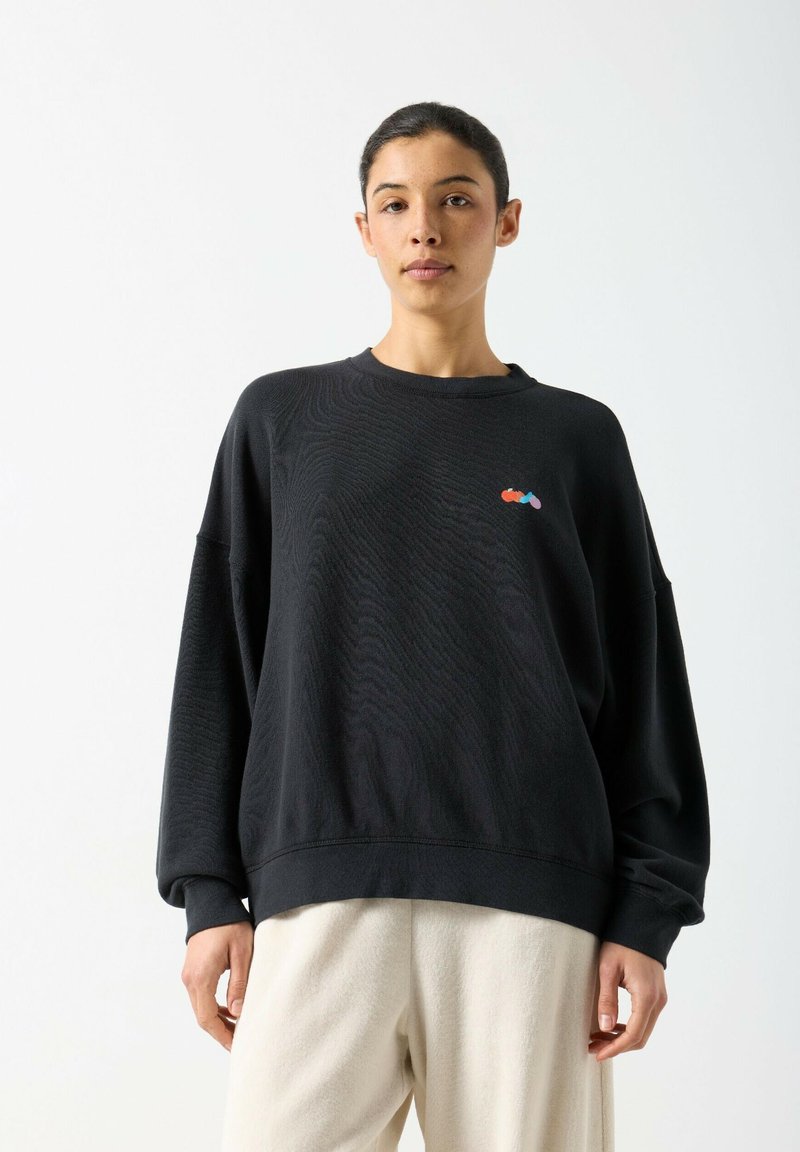 Woman wearing an oversized black sweatshirt with small colorful embroidery on the chest and loose light beige pants, standing against white background.
