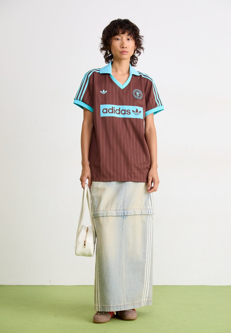 Jersey marrón con rayas verticales y acentos turquesa, acompañado de una falda maxi de mezclilla azul claro, sosteniendo un pequeño bolso blanco.