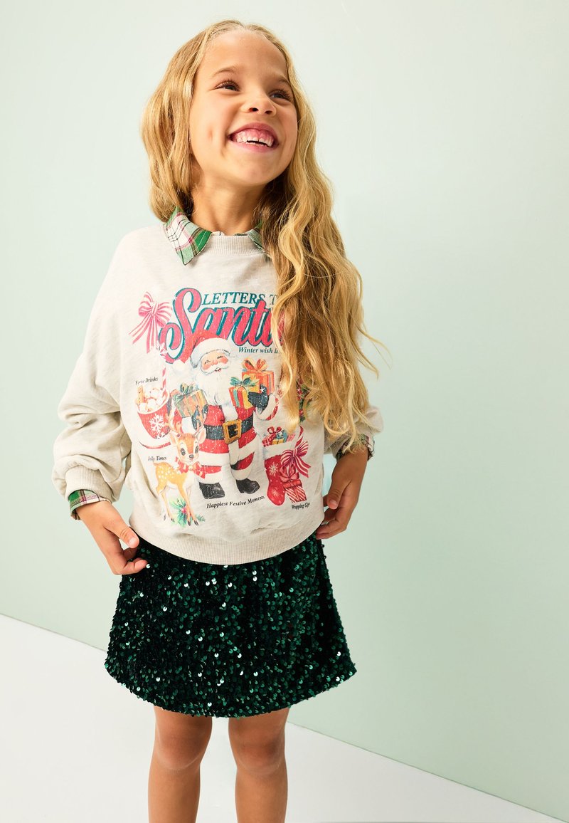 Sweat-shirt gris clair avec un motif festif de Père Noël, associé à une jupe à sequins verts. La tenue comprend également un col de chemise à carreaux visible en dessous.