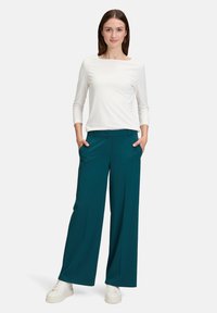 Haut à manches longues blanc avec un décolleté carré associé à un pantalon ample teal, doté de poches latérales et d'un tissu texturé, porté avec des baskets blanches.