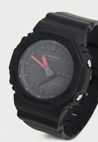 Montre G-SHOCK noire avec un cadran rond, affichage numérique, accents rouges, bracelet en caoutchouc et boîtier texturé. Les caractéristiques incluent une résistance à l'eau et plusieurs boutons.
