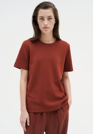 Rostfarbener Kurzarm-T-Shirt aus einem glatten, weichen Stoff mit Rundhalsausschnitt und lässiger Passform, getragen mit passenden Hosen.