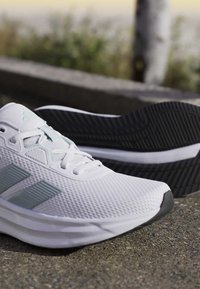 Chaussures de sport blanches avec des bandes grises, dessus en mesh texturé et semelles noires à crampons reposant sur un pavé extérieur.