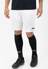 Weiße Fußballshorts mit einem schwarzen Logo, kombiniert mit schwarzen knielangen Socken mit einem weißen Logo und schwarzen Schienbeinschonern mit strukturiertem Design.