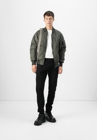 Olivenfarget bomberjakke med ribbede mansjetter og krage, hvit t-skjorte, svarte slim-fit jeans og svarte kraftige sko med tykk såle. Moderat tekstur.