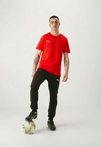 Tânăr îmbrăcat într-un tricou roșu Puma și pantaloni negri, echilibrând o minge de fotbal colorată cu piciorul drept pe un fundal alb simplu.
