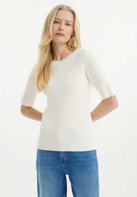 Bílý žebrovaný pletený top s krátkými rukávy, který má kulatý výstřih a knoflíkové detaily na manžetách rukávů. Kombinováno s modrými džíny.