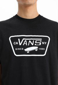 Svart t-shirt i bomull, med en vit grafisk logotyp som inkluderar "VANS", en skateboardikon och texten "SEDAN 1966".