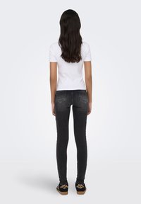 Γυναίκα με μακριά σκούρα μαλλιά, φοράει λευκό κοντομάνικο πουκάμισο, μαύρα στενά τζιν και μαύρα παπούτσια με λεπτομέρειες λεοπάρ, στέκεται με την πλάτη στραμμένη.