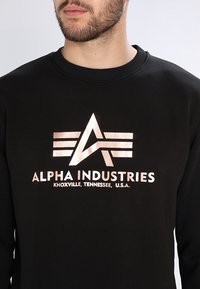 Mies, joka käyttää mustaa hupparia, jossa on metallinen "Alpha Industries Knoxville, Tennessee, Yhdysvallat" -logo ja emblem rintataskussa.