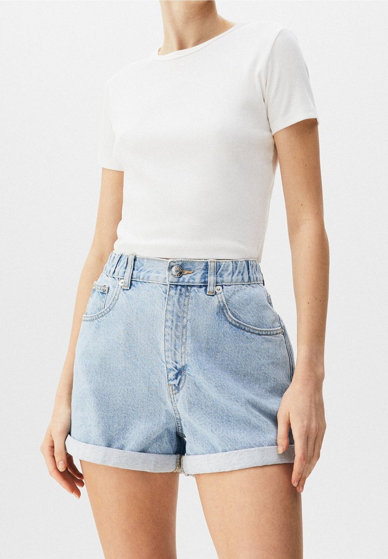 Femme portant un haut blanc à manches courtes côtelé et un short en denim bleu clair taille haute avec ourlet roulé, debout devant un fond uni.