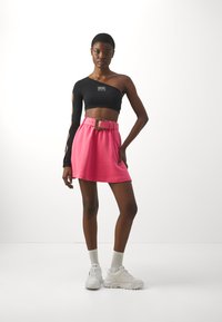 Svart en-skulder crop top med utskårne ermer, parret med et rosa belted skjørt med en gullfarget spenne. Bruges med hvite sneakers.