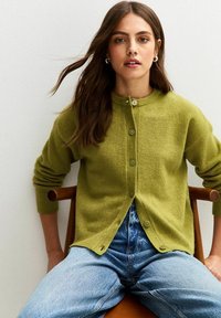 Groene wollen cardigan met ronde hals en knoopsluiting, met geribde mouwen en zoom. Draagt over lichtblauwe denim jeans.