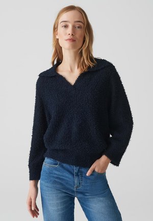 Marineblauer Pullover mit einer strukturierten, samtigen Oberfläche, tiefem V-Ausschnitt und weitem Kragen. Kombiniert mit hellblauen Jeans.