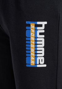 Melni sporta bikses ar lielu, izteiksmīgu „hummel” logotipu baltā, zilā un oranžā krāsā. Audums izskatās mīksts un teksturēts, ideāli piemērots sporta apģērbam.