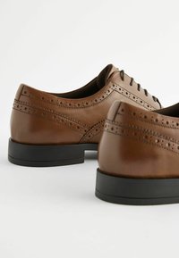 Pantofi de dress din piele maro, cu detalii perforate, o textură netedă, vârful rotund și tălpi din cauciuc negru. Șireturile sunt vizibile în față.