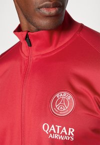 Red athletic zip jacket featuring a stand collar, embroidered Paris Saint-Germain logo, and Qatar Airways branding in white text. Smooth texture.