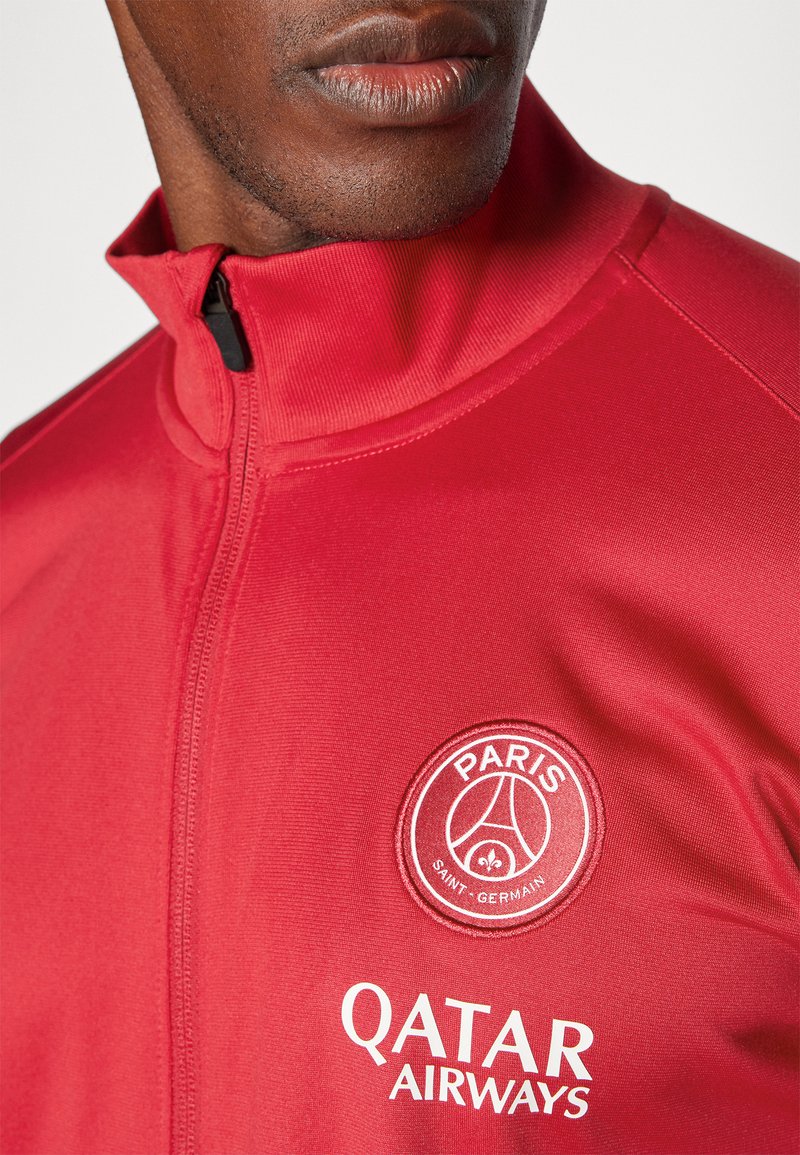 Chaqueta deportiva roja con cremallera, que presenta un cuello alzado, el logo bordado del Paris Saint-Germain y el branding de Qatar Airways en texto blanco. Textura suave.