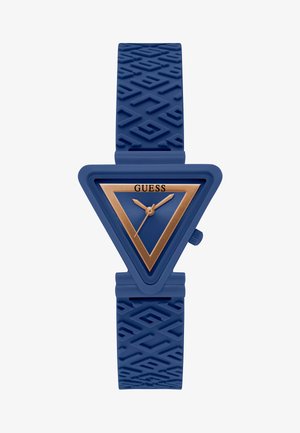 Montre en caoutchouc bleue avec un boîtier triangulaire présentant un cadran triangulaire doré et un bracelet texturé bleu. Nom de la marque "GUESS" affiché.