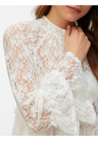 Hvit bluse i blonde med høy hals, lange ermer og volangmansjetter. Har blomsterdekor og et gjennomsiktig, teksturert stoff.