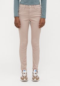 Jeans ajustados rosa claro con una textura suave, diseño de cinco bolsillos y herrajes plateados; usados con zapatillas deportivas blancas y beige.