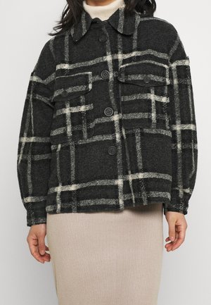 Femme portant une veste à carreaux noirs et blancs à boutons par-dessus une jupe en maille côtelée beige, visible des épaules jusqu'aux hanches.