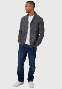Cardigan grigio lavorato a maglia con collo a scialle, chiusura a bottoni, tessuto testurizzato, indossato sopra una maglietta bianca e abbinato a jeans blu e sneaker bianche.