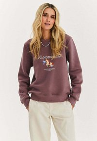 Fioletowa bluza z okrągłym dekoltem, z grafiką jednorożca i napisem. Miękka tkanina, ściągacze na mankietach i dole. Połączona z kremowymi spodniami.