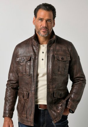 Mann trägt eine dunkelbraune Lederjacke über einem weißen Hemd mit Knöpfen und steht mit einer Hand in der Tasche vor einem einfarbigen hellen Hintergrund.