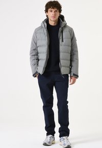 Chaqueta acolchada gris con capucha, que presenta un cierre con cremallera en la parte delantera, mangas texturizadas y costuras en ángulo. Llevada sobre un suéter de punto azul oscuro y pantalones azul marino.