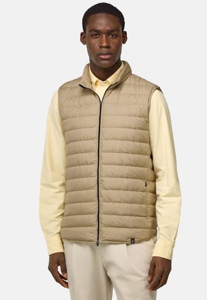 Gilet rembourré beige avec fermeture éclair à l'avant et col haut, présentant des coutures horizontales, associé à une chemise à manches longues jaune clair.