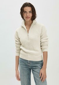 Helles beiges Strickpullover mit halbem Reißverschlusskragen, gerippter Textur und enganliegenden Ärmeln; kombiniert mit blauen Jeans.