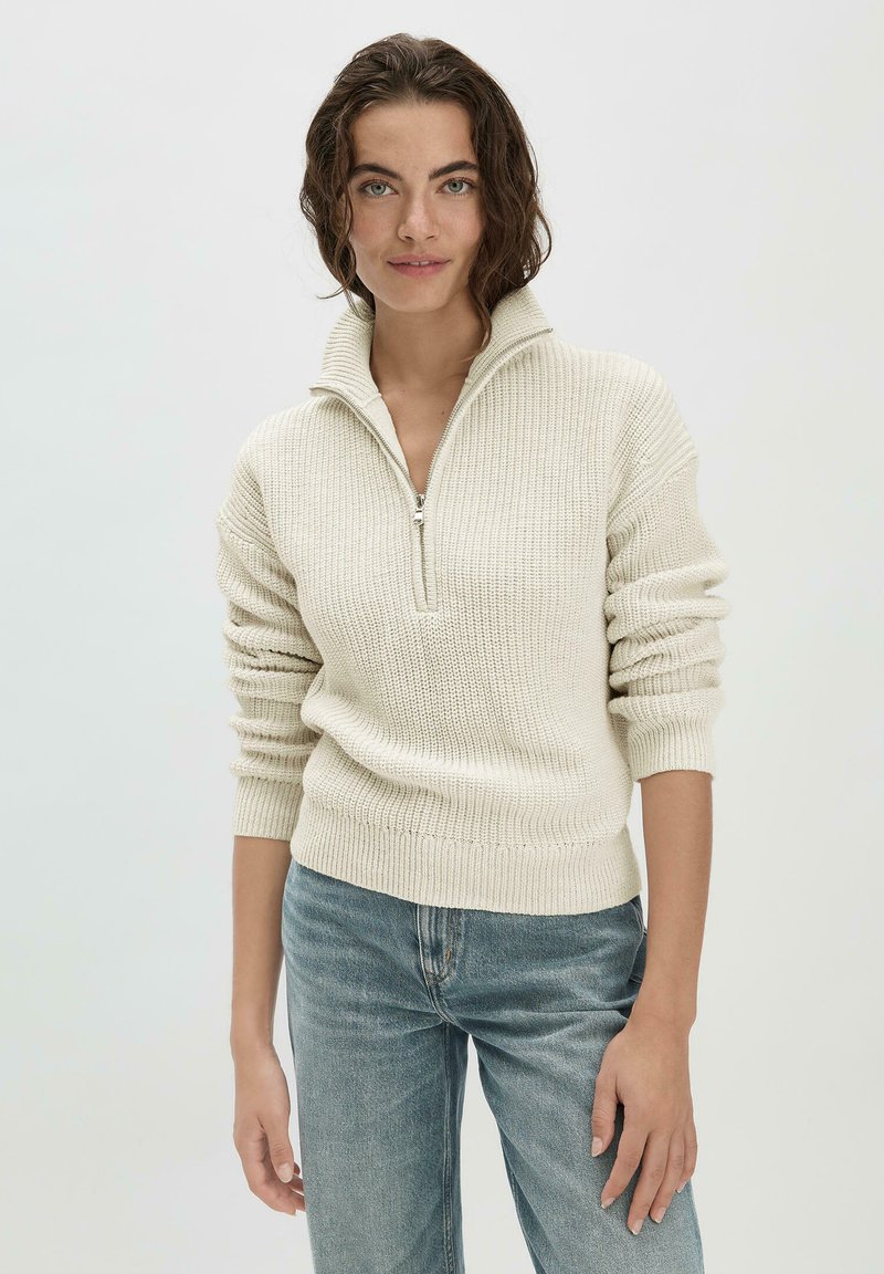 Helles beiges Strickpullover mit halbem Reißverschlusskragen, gerippter Textur und enganliegenden Ärmeln; kombiniert mit blauen Jeans.