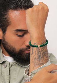 Grünes Perlenarmband mit goldenen Akzenten, das unterschiedliche Perlenformen und -größen aufweist, getragen an einem Handgelenk mit sichtbaren Tattoos.