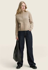 Beige fleece zip-up jacket with a round neckline and front pocket, paired with dark blue wide-leg jeans and a black tote bag.