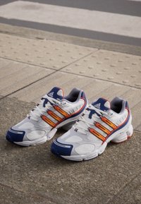 Bijele atletske tenisice s narančastim i plavim detaljima, s mrežastim panelima, dizajnom s tri pruge i jastučićima u sole.