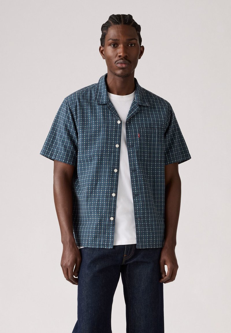 Korte mouwen knoopshirt in donkerblauw, met een ruitpatroon en lichtblauwe accenten. Gemaakt van lichte stof, gecombineerd met een wit T-shirt.