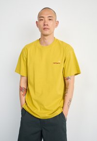 Geel katoenen t-shirt met een ronde halslijn en korte mouwen, met een klein oranje logo op de linkerkant van de borst. Gecombineerd met donkere shorts.