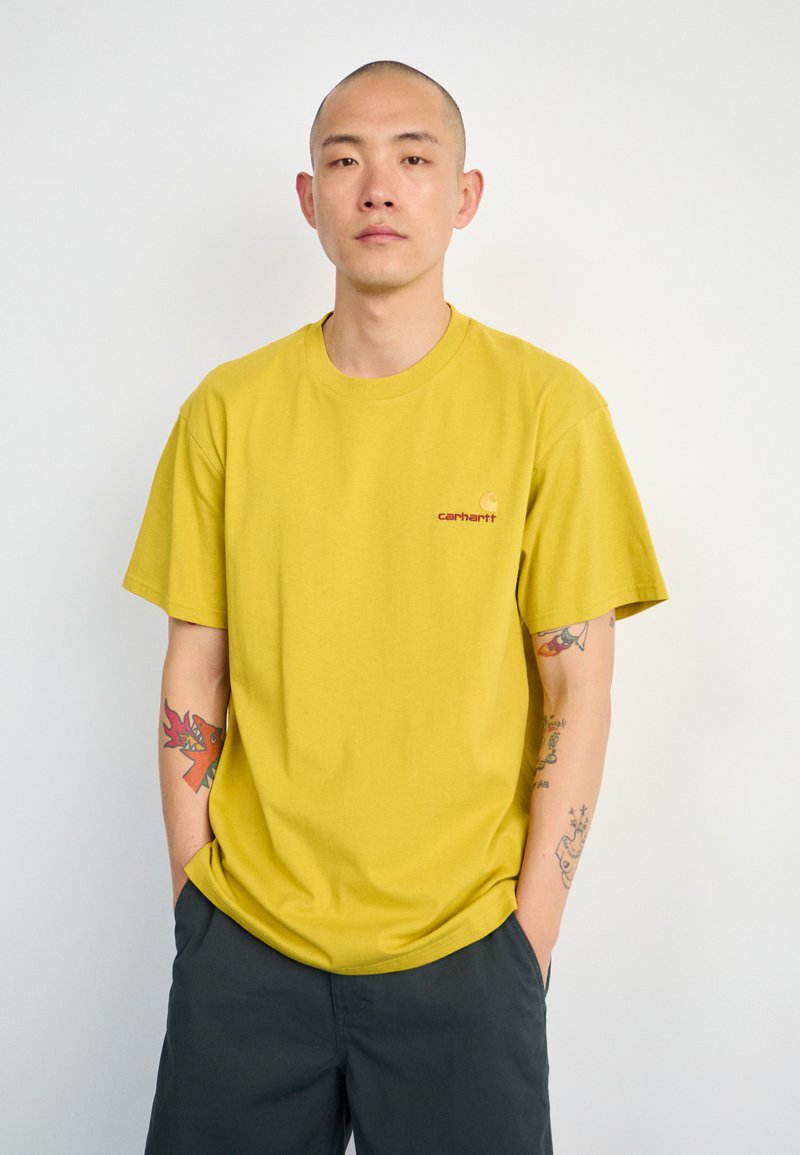 Geel katoenen t-shirt met een ronde halslijn en korte mouwen, met een klein oranje logo op de linkerkant van de borst. Gecombineerd met donkere shorts.