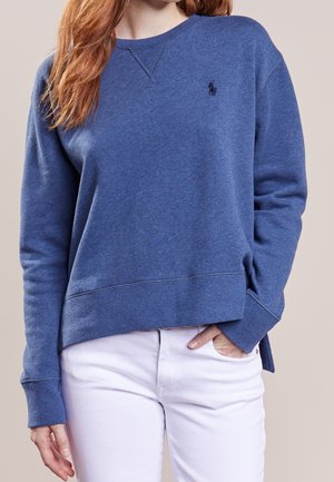 Femme portant un sweat-shirt bleu à manches longues avec un petit logo et un pantalon blanc, une main dans la poche et l'autre détendue.