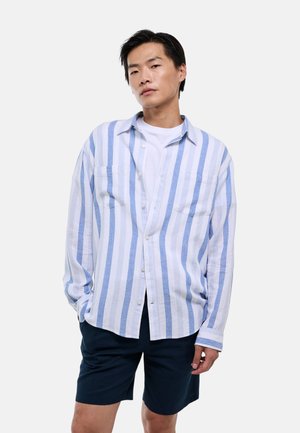 Homme portant une chemise à rayures verticales blanches et bleues par-dessus un t-shirt blanc et un short bleu foncé, debout avec une main dans la poche.