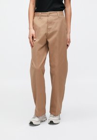 Φόρμες chino από βαμβάκι σε καφέ απόχρωση με ευθεία γραμμή, διαθέτουν κουμπωτό κλείσιμο και πλαϊνές τσέπες. Φοριούνται με αθλητικά παπούτσια ανοιχτού χρώματος.