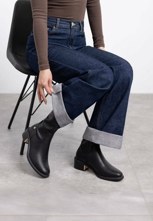 Stivaletti in pelle nera con tacco basso e dettagli dorati, abbinati a jeans blu scuro a vita alta e una maglietta rigida marrone, seduta su una sedia.