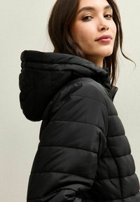 Giacca a vento nera con cappuccio, caratterizzata da una texture trapuntata e cuciture orizzontali, chiusura lampo robusta e una silhouette aderente.