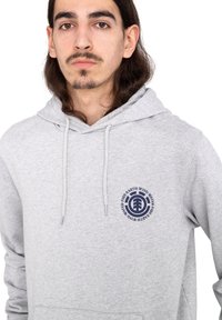 Sudadera ligera gris con cordones, que presenta un logotipo circular en azul marino en el pecho y un bolsillo frontal. Textura de tejido suave, diseño informal.