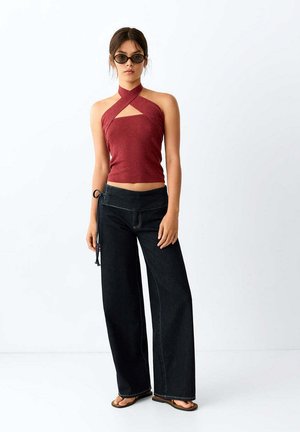 Mujer con top burdeos de cuello halter, vaqueros negros de pierna ancha con lazada lateral, gafas de sol negras y sandalias marrones, de pie frente a un fondo blanco.