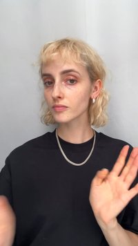 En person med kort, vågigt blont hår bär en svart t-shirt. Utmärkande drag inkluderar silverfärgade ringörhängen och ett silverfärgat nät-halsband.