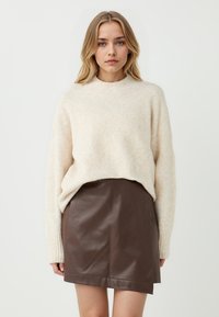 Maglione a maglia beige chiaro con polsini a costine, abbinato a una minigonna in pelle marrone. L'outfit presenta una vestibilità rilassata e texture morbide.